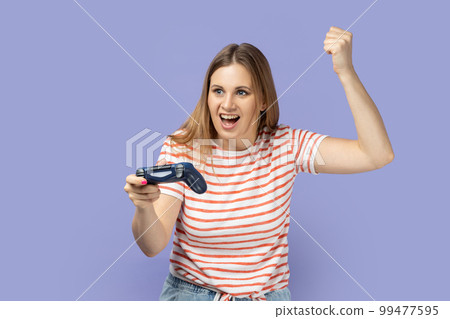 Portrait of excited amazed blond woman wearing striped T-shirt holding in hands gamepad joystick, clenched fist, showing yes gesture, winning. Indoor studio shot isolated on purple background. 99477595