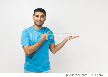 Portrait of handsome joyful smiling happy unshaven man wearing blue T- shirt standing presenting advertisement area on his palm, mockup. Indoor studio shot isolated on gray background. 99477670