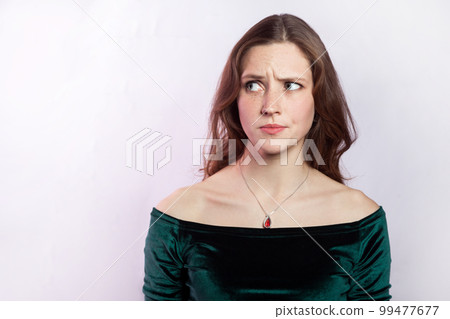 Portrait of confused puzzled pensive woman wearing green dress looking away with pensive thoughtful expression, thinking about problem. Indoor studio shot isolated on gray background. 99477677