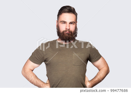 Portrait of bearded man wearing dark green T-shirt looking cross-eyed with stupid facial expression, fooling around, keeps hands on hips. Indoor studio shot isolated on light gray background. 99477686