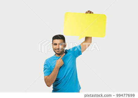 Portrait of optimistic smiling unshaven man wearing blue T- shirt standing pointing at yellow speech frame, space for words and thoughts. Indoor studio shot isolated on gray background. Portrait of optimistic smiling unshaven man wearing blue T- shirt standing pointing at yellow speech frame, space for words and thoughts. Indoor studio shot isolated on gray background. 99477699