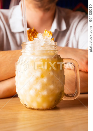 Pina colada cocktail with ice, pineapple chips, cocktail straw in glass with handle on wooden table, male man hands. Front view, close-up, vertical photo. 99480010