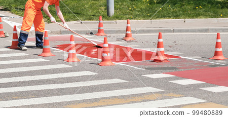 Road repair. Man in orange work clothes repairs pedestrian crossing in morning, summer and spring. Road is fenced with special cones. Road repair. Man in orange work clothes repairs pedestrian crossing in morning, summer and spring. Road is fenced with special cones. 99480889