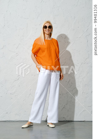 An adult woman posing in a stylish orange blouse and white trousers 99481316