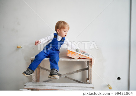 Cute kid construction worker sitting on wooden table against white wall in apartment under renovation. Cheerful little boy wearing safety helmet and work overalls while playing at home. Cute kid construction worker sitting on wooden table against white wall in apartment under renovation. Cheerful little boy wearing safety helmet and work overalls while playing at home. 99481864