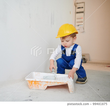 Adorable child construction worker crouching down and preparing paint roller for wall painting. Kid wearing safety helmet and work overalls while working on home renovation. 99481875