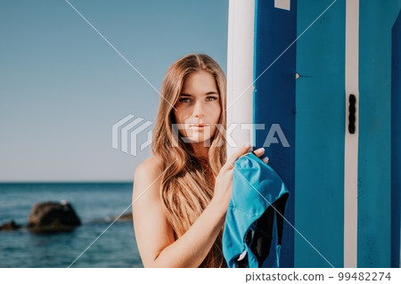 Woman sea sup. Close up portrait of happy young caucasian woman with long hair looking at camera and smiling. Cute woman portrait in a blue bikini posing on sup board in the sea 99482274