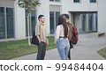 Group of positive multiracial students in casual clothes looking at each other and discussing project while standing near university building with folders and bags 99484404