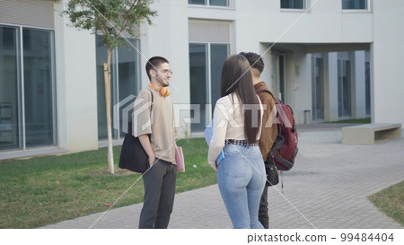 Group of positive multiracial students in casual clothes looking at each other and discussing project while standing near university building with folders and bags Group of positive multiracial students in casual clothes looking at each other and discussing project while standing near university building with folders and bags 99484404