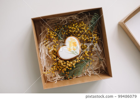 Easter gingerbread cookie in shape of a heart in a gift box with colorful yellow mimosa flowers. Eastet, Spring, April card or banner. Top view Easter gingerbread cookie in shape of a heart in a gift box with colorful yellow mimosa flowers. Eastet, Spring, April card or banner. Top view 99486079
