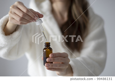 Young woman in a bathrobe is holding a bottle with a dropper of oil or serum for face and body care. 99486131