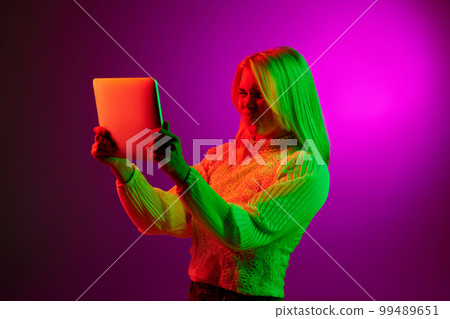 Communication. Emotive young woman having online video call on tablet over magenta studio background in green neon light. Concept of emotions, lifestyle 99489651