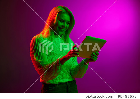 Modern technologies, online services. Smiling young woman looking on tablet over magenta studio background in green neon light. Concept of emotions, lifestyle 99489653