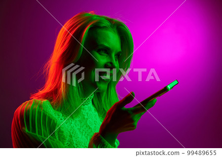 Happy smiling young woman talking on mobile phone, recording voice messages over magenta studio background in green neon light. Concept of emotions, lifestyle 99489655