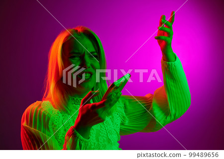 Young woman emotionally recording voice message on mobile phone over magenta studio background in green neon light. Concept of emotions, lifestyle 99489656
