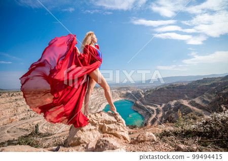 woman red dress lake mountains. Side view of a woman in a long red dress posing on a rock high above the lake. Against the background of the blue sky and the lake in the form of a heart 99494415