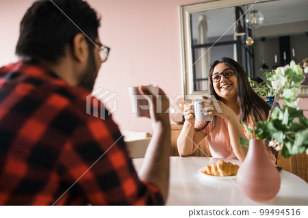 Happy couple eating breakfast and talking at dining table in morning. Indian girl and latin guy. Relationship and diversity concept Happy couple eating breakfast and talking at dining table in morning. Indian girl and latin guy. Relationship and diversity concept 99494516