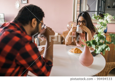 Happy couple eating breakfast and talking at dining table in morning. Indian girl and latin guy. Relationship and diversity concept Happy couple eating breakfast and talking at dining table in morning. Indian girl and latin guy. Relationship and diversity concept 99494533