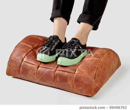Female legs in sneakers resting on terracotta leather footrest 99496762