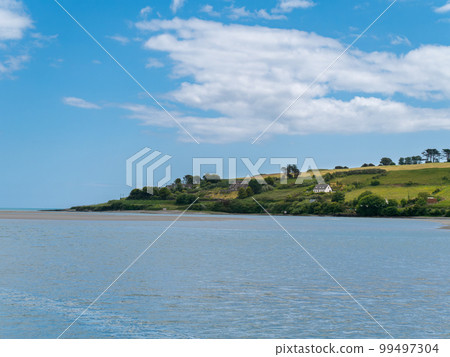 Beautiful sky over a water surface on a summer day. The picturesque green coast of Ireland. Several buildings on the hill. Seascape. Green trees on island Beautiful sky over a water surface on a summer day. The picturesque green coast of Ireland. Several buildings on the hill. Seascape. Green trees on island 99497304