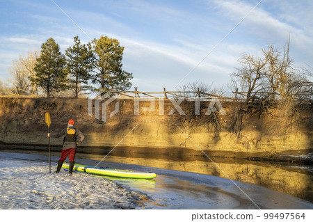 male paddler with inflatable stand up paddleboard on an icy shore of the Poudre River in Fort Collins, Colorado 99497564