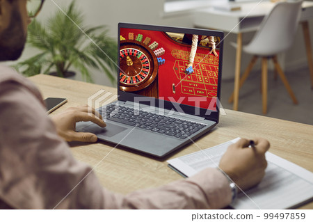 Man placing bets and writing notes while playing online casino at home on his laptop. 99497859