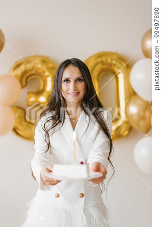 Positive woman celebrating anniversary dressed in stylish feather dress posing with birthday candle cake around inflated balloons at home 99497890