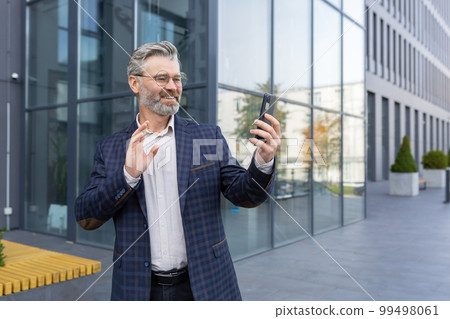 Successful mature man in business suit using app on phone for video call, senior businessman outside office building talking remotely with friends waving at smartphone camera. Successful mature man in business suit using app on phone for video call, senior businessman outside office building talking remotely with friends waving at smartphone camera. 99498061