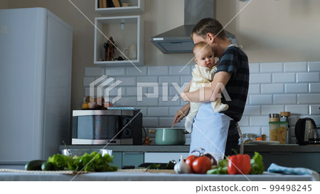 Young father in apron holding cute baby in hands while cooking dinner 99498245