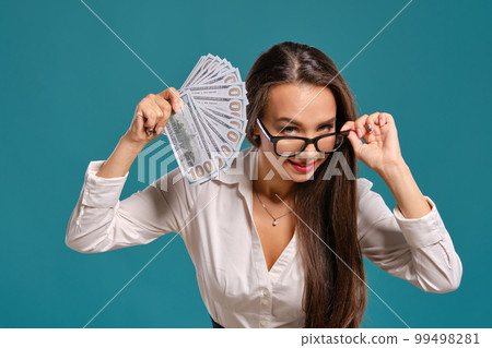 Brunette girl in glasses, wearing in a black short skirt and white blouse is posing holding a fan of hundred dollar bills against a blue background. 99498281
