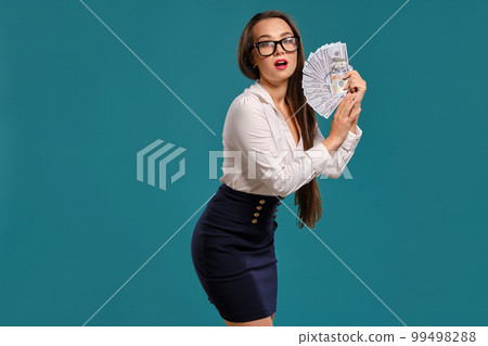 Brunette girl in glasses, wearing in a black short skirt and white blouse is posing holding a fan of hundred dollar bills against a blue background. Brunette girl in glasses, wearing in a black short skirt and white blouse is posing holding a fan of hundred dollar bills against a blue background. 99498288