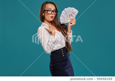 Brunette girl in glasses, wearing in a black short skirt and white blouse is posing holding a fan of hundred dollar bills against a blue background. 99498289