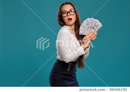 Brunette girl in glasses, wearing in a black short skirt and white blouse is posing holding a fan of hundred dollar bills against a blue background. Brunette girl in glasses, wearing in a black short skirt and white blouse is posing holding a fan of hundred dollar bills against a blue background. 99498290