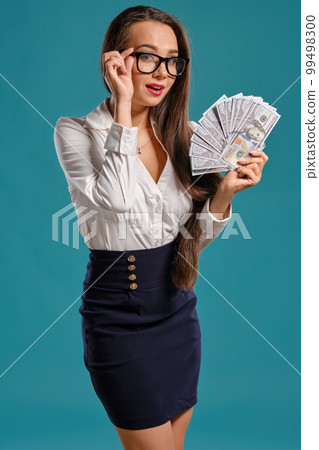 Brunette girl in glasses, wearing in a black short skirt and white blouse is posing holding a fan of hundred dollar bills against a blue background. 99498300