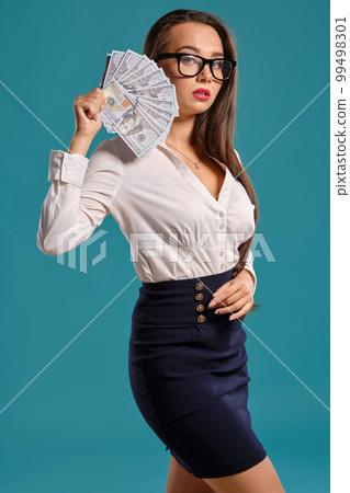Brunette girl in glasses, wearing in a black short skirt and white blouse is posing holding a fan of hundred dollar bills against a blue background. Brunette girl in glasses, wearing in a black short skirt and white blouse is posing holding a fan of hundred dollar bills against a blue background. 99498301