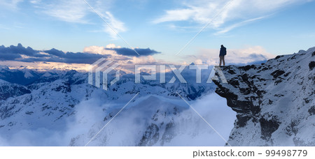 Adventurous Man Hiker standing on top of icy peak with rocky mountains in background. 99498779