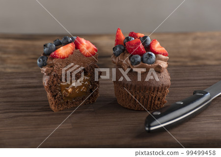 Cut chocolate cupcake is decorated with chocolate butter cream and berries. Dessert and knife on rough wooden brown board. Cut chocolate cupcake is decorated with chocolate butter cream and berries. Dessert and knife on rough wooden brown board. 99499458