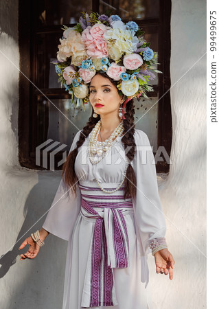 Brunette girl in a white ukrainian authentic national costume and a wreath of flowers is posing against a white hut. Close-up. 99499675