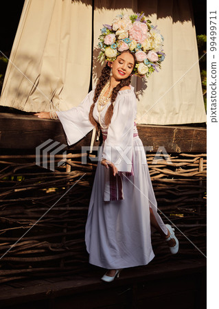 Brunette girl in a white ukrainian authentic national costume and a wreath of flowers is posing against a terrace. Brunette girl in a white ukrainian authentic national costume and a wreath of flowers is posing against a terrace. 99499711