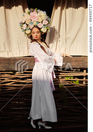 Brunette girl in a white ukrainian authentic national costume and a wreath of flowers is posing against a terrace. 99499717