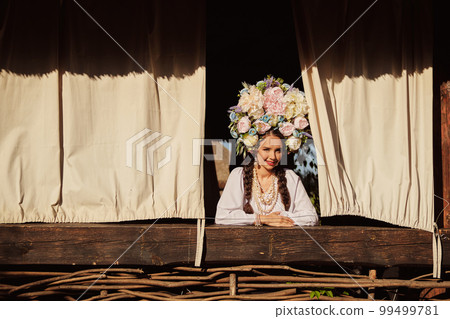 Brunette girl in a white ukrainian authentic national costume and a wreath of flowers is is looking out of the window. Brunette girl in a white ukrainian authentic national costume and a wreath of flowers is is looking out of the window. 99499781