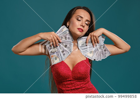 Brunette girl with a long hair, wearing a sexy red dress is posing holding a fan of hundred dollar bills against a blue background. Brunette girl with a long hair, wearing a sexy red dress is posing holding a fan of hundred dollar bills against a blue background. 99499858