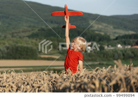 Child boy in red T-shirt plays with toy plane in wheat field on mountains background. Concept of flights and travel with children. 99499894