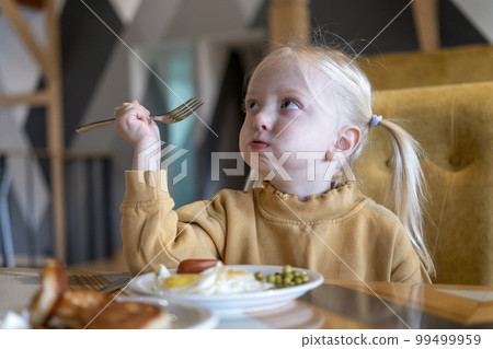 Fair-haired girl with two ponytails sits at table holding fork and has breakfast. Mouth full of food. Little girl eats 99499959
