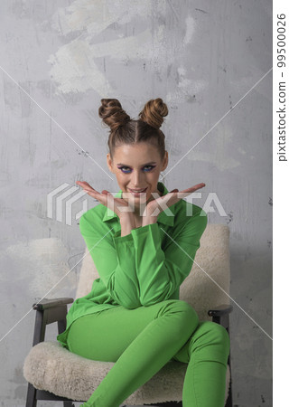 Bright girl with evening makeup in green pantsuit sits on chair on gray wall background. Vertical frame Bright girl with evening makeup in green pantsuit sits on chair on gray wall background. Vertical frame 99500026