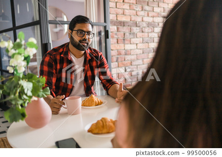 Young diverse loving couple eating croissant and talks together at home in breakfast time. Communication and relationship concept 99500056