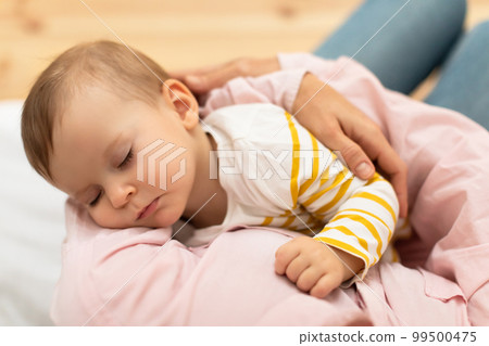 Loving mom carying of her baby daughter at home, mother holding sleeping infant child on hands, above view 99500475