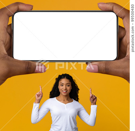 Happy Black Woman Pointing Up At Big Blank Smartphone In Giant Hands 99500480