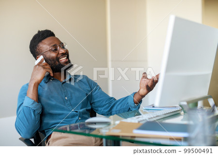 Excited black businessman talking on cellphone and looking at computer monitor, sitting at workplace in office Excited black businessman talking on cellphone and looking at computer monitor, sitting at workplace in office 99500491