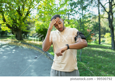 Young African American man runner, athlete in the park. A tired man counts his pulse on a fitness bracelet. Surprised, he looks at the result on the smartwatch on his hand. Wipes sweat with his hand. Young African American man runner, athlete in the park. A tired man counts his pulse on a fitness bracelet. Surprised, he looks at the result on the smartwatch on his hand. Wipes sweat with his hand. 99500792